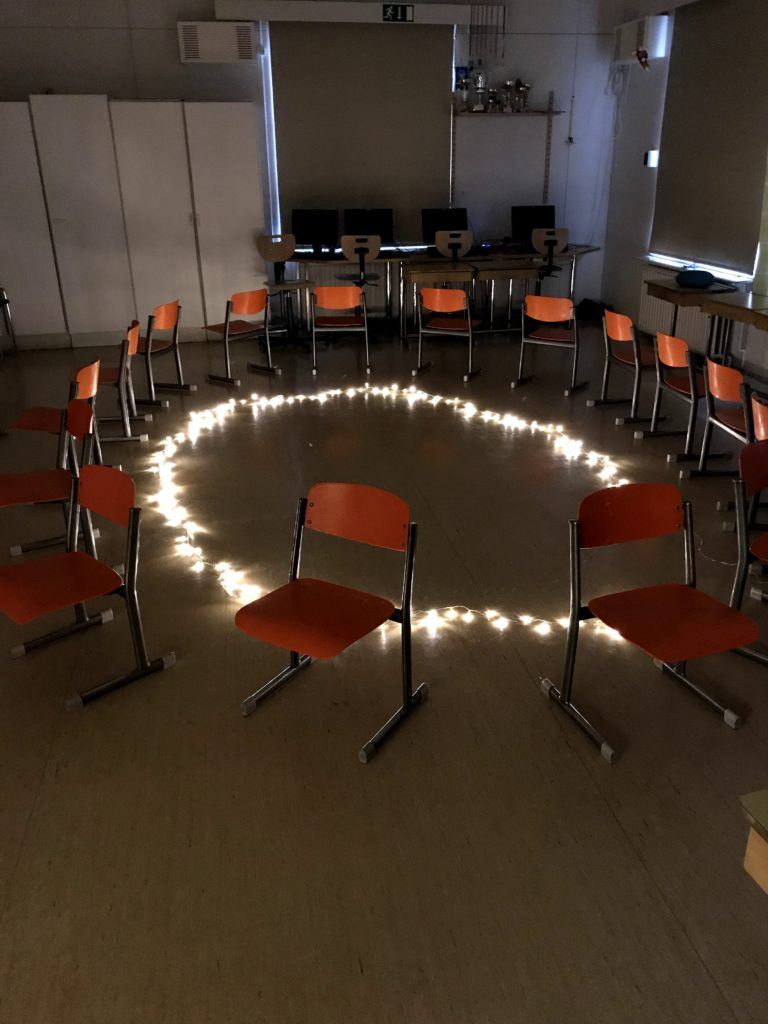 Koululuokassa tuoleja asetettuna ympyrän muotoon selkänojat ympyrän keskustaan. Luokassa on tunnelmallinen valaistus.