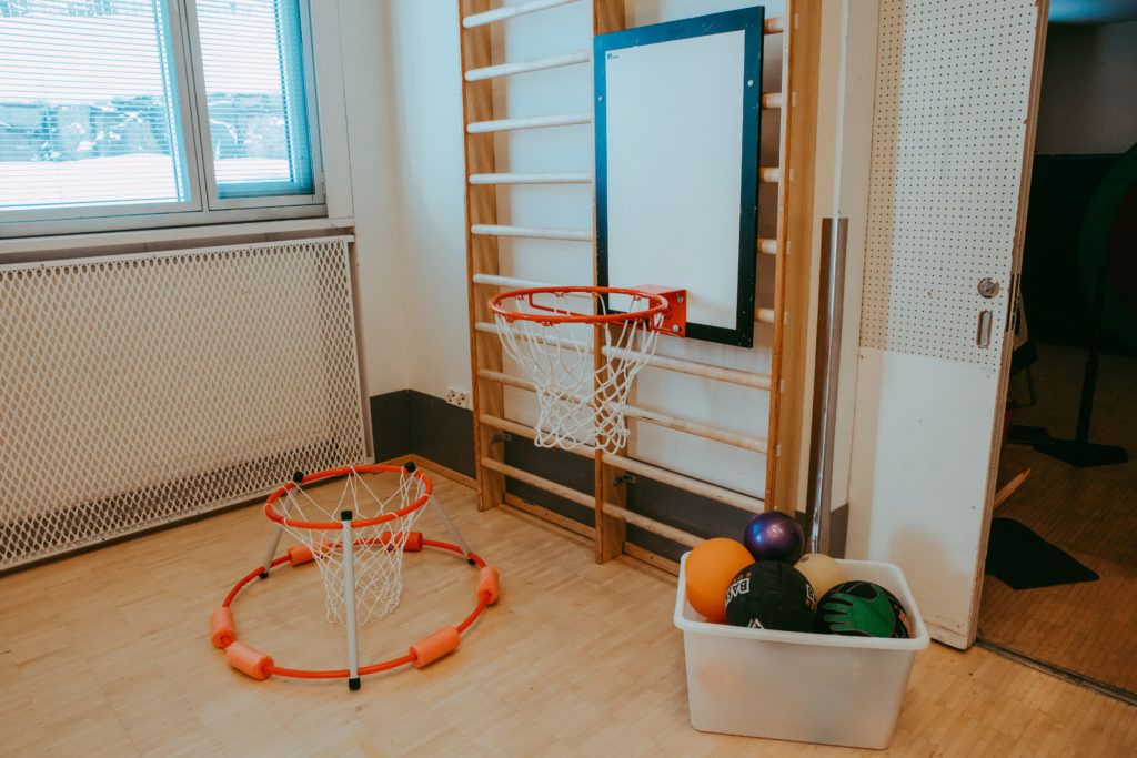 Liikuntasalin puolapuihin kiinnitetty koripallokori. Lattialla koripallokori ja laatikollinen erilaisia palloja.
